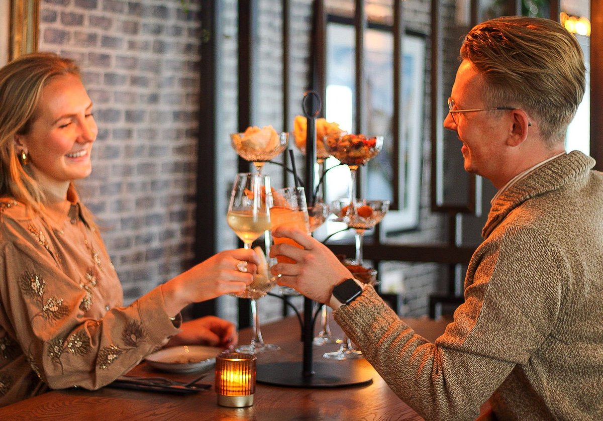 Twee gasten proosten met een glas wijn en een cocktail, met een Taste Tree vol kleine gerechtjes en een brandende kaars op tafel